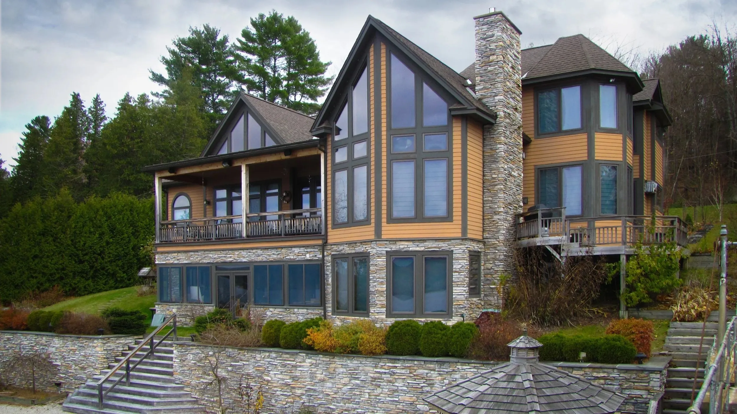 Custom lakeside home with a tall wall of gable windows, a stone chimney, and two levels of decks